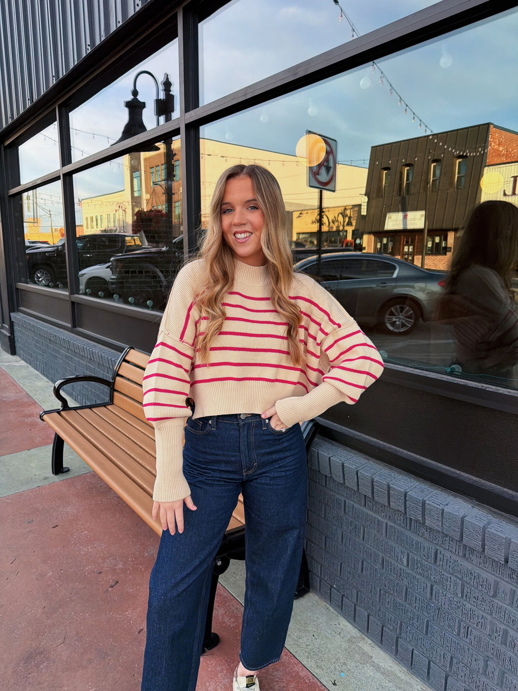 Woman in a striped sweater and jeans standing on a sidewalk with a building and street in the background.